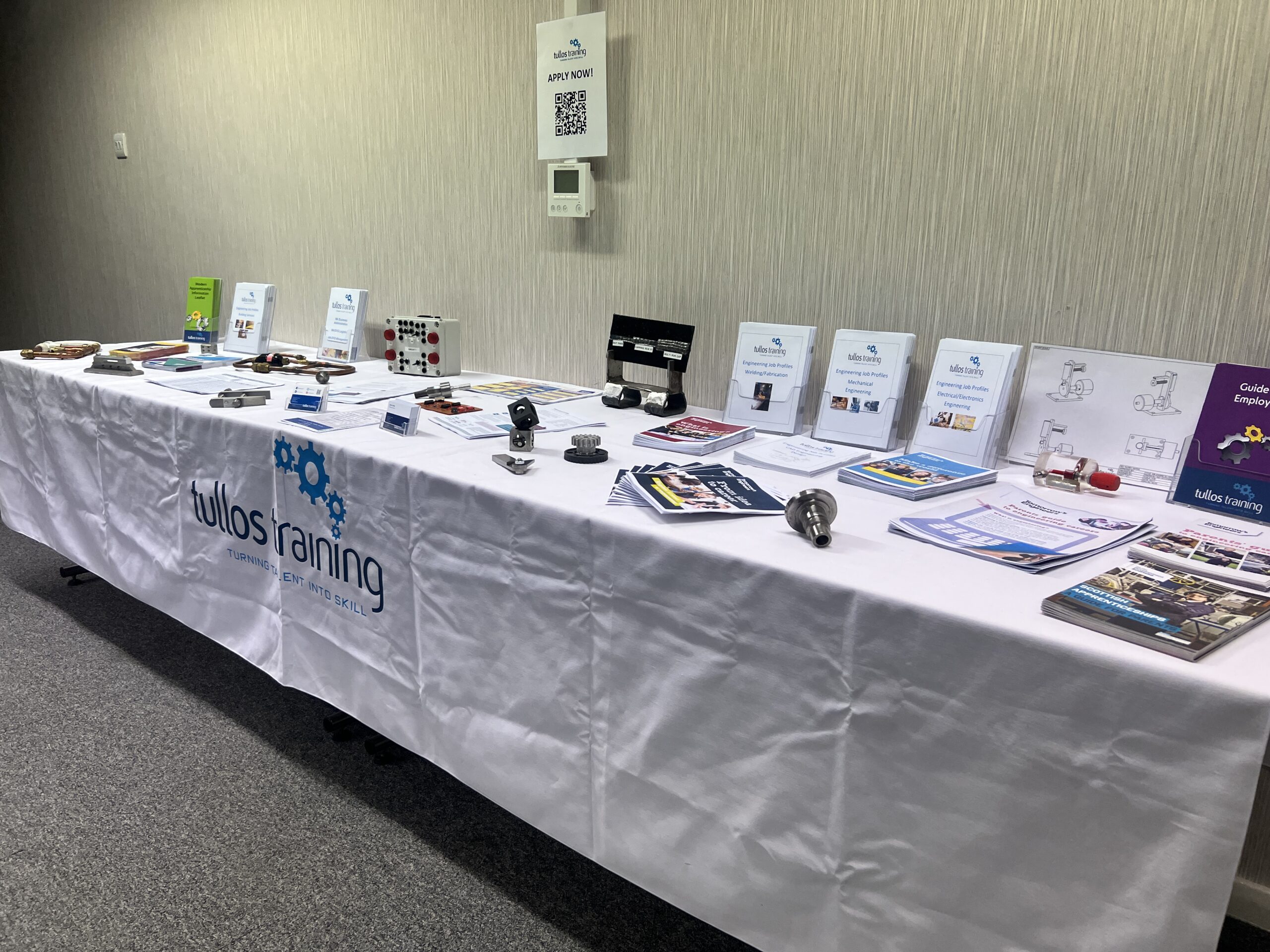 Image shows Tullos Training's careers fair table set up with a white branded table cloth.  Informational leaflets are displayed on the table along with example pieces from their engineering related and plumbing workshops.